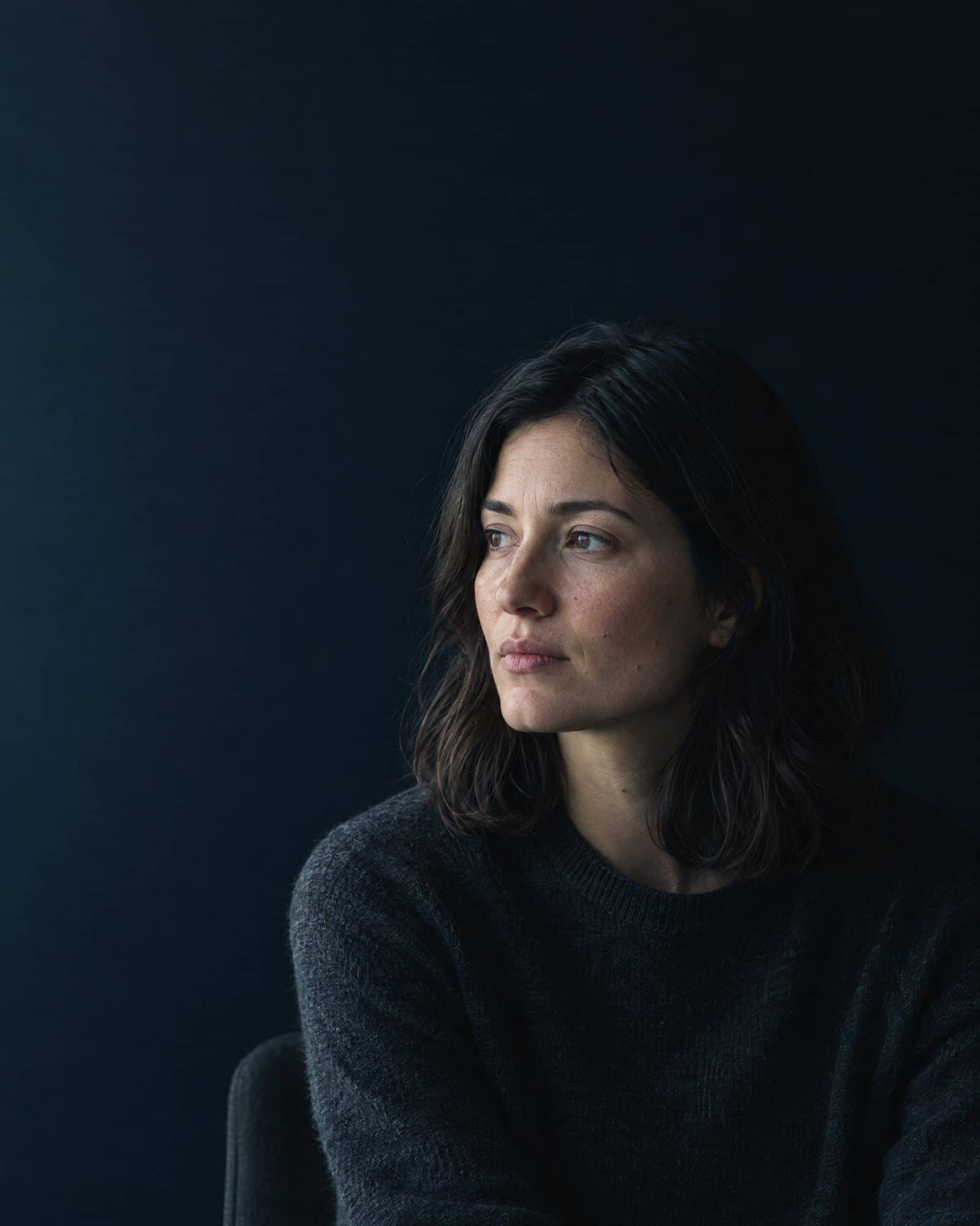 AI-generated portrait of a woman in a dark sweater against a dark blue wall, looking away from the camera. She is not a real person.